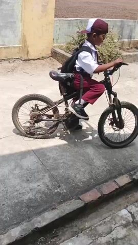 A young person wearing a white shirt, maroon pants, and a white and maroon baseball cap is riding a bicycle. They are carrying a black backpack. The bicycle appears to be a mountain bike with knobby tires and a front suspension. The rider is moving from left to right across the frame. The background shows a textured wall on the left and a dirt area with some greenery behind the rider. The ground is paved with concrete. The lighting suggests it is daytime and sunny.