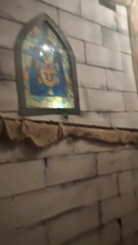 The video shows a man walking down a hallway. The hallway is decorated to look like a dungeon, with faux stone walls. There are also decorations like stained glass windows, banners, and string lights. There are AC units on the wall. The man is wearing a black jacket.