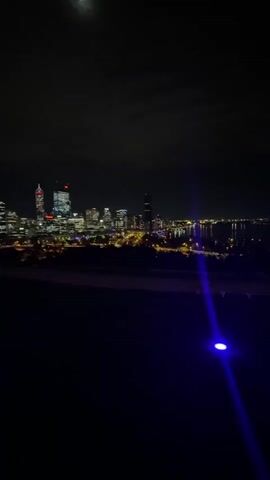 The video depicts a night scene with a distant city skyline illuminated against a dark sky with a moon. The foreground is dark, but a ground-level light shines, shifting from blue to red. There are no visible people or actions in the video.