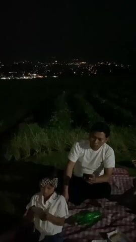 A man and a young girl are picnicking at night in an outdoor setting, with a view of a city skyline in the distance. The man, wearing a white t-shirt and dark pants, is seated on a red and white checkered blanket, holding a smartphone. The girl, in a white shirt and dark pants, sits beside him, also looking at a smartphone and holding a white object. A green bottle and a food container are visible on the blanket. The background features rows of plants, likely crops, and beyond that, the twinkling lights of a city spread across hills. The sky is dark, indicating nighttime. The overall mood appears relaxed and informal, suggesting a family outing or a quiet evening spent together. The location is identified as Kesiman, Indonesia.