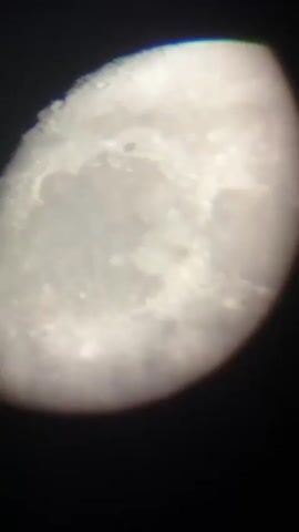 The video shows a close-up, slightly blurry view of the moon. The moon is partially illuminated, with a portion appearing bright and detailed, showing craters and surface features. The edges of the moon are rounded and illuminated, while the surrounding area is dark. There is a subtle shift in brightness and focus throughout the short clip, suggesting slight movement or adjustments to the camera or telescope.