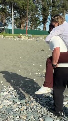 Two people are on a beach with dark sand and some grey stones. One person is giving the other a piggyback ride. The person carrying is wearing black slacks and white sneakers. The person being carried is wearing reddish brown pants and a white shirt. Trees are visible in the background. The sky is bright.