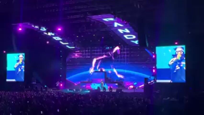 The video shows a live concert with a large crowd facing a stage. The stage features large screens displaying images of the performer, Randy. The screens flank the central stage area where the performance is taking place. Stage lights and a banner with "Randy" are visible above the stage. The crowd appears to be enjoying the performance, with many people standing and watching the show.