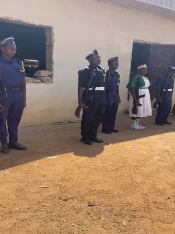 Five young individuals stand in a disciplined line on a patch of dry, sandy ground, bathed in bright sunlight. Behind them is a simple, light-colored, single-story building with several open window and door frames, some showing exposed brick or stone around the edges.

Four of the individuals are male, dressed in dark blue uniforms consisting of trousers, jackets with light blue shirts and dark ties, and dark peaked caps. They wear belts with pouches and shoulder straps. Each holds a wooden prop, possibly an imitation rifle, vertically at their side. The fifth individual, positioned near the center, is female. She wears a white dress, a green sleeveless top or vest, and white socks with dark shoes. She also holds a similar wooden prop and wears a golden crown on her head.

The strong shadows cast on the ground indicate it is a clear, sunny day. The individuals maintain a formal and attentive stance, suggesting they are participating in a drill, parade, or ceremonial event. The setting appears to be outdoors, in what looks like a community or school compound in Andaha, Nigeria. There is no visible text in the image.