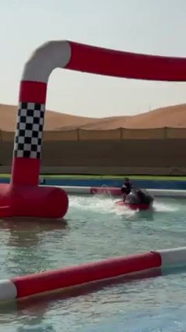 The video shows a person driving a small, black and red watercraft on a pool with inflatable red and white barriers. The person is wearing a black helmet and clothes. The pool is located in a desert-like environment with sand dunes and sparse vegetation. The watercraft is making waves as it moves through the water, maneuvering around the barriers. The background shows a light sky, suggesting it is daytime.