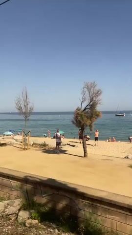 The video shows a scenic view of the coastline in El Masnou, Spain. The time of day appears to be late morning or early afternoon, with clear, sunny skies and calm waters. The scene transitions from a beach with people relaxing under umbrellas to a walkway along the sea.

In the initial frames, several individuals are visible on the sandy beach, some are swimming in the ocean, while others are seated or standing near colorful beach umbrellas. A person on an electric scooter, wearing a helmet and backpack, rides along the paved pathway bordering the beach. A tall lamppost stands near the walkway, with a sign attached to it. A small boat is visible in the distance on the water.

As the video progresses, the focus shifts to different sections of the coastline. The walkway, made of yellow-toned paving stones, runs alongside a low, brick wall adorned with graffiti. Rocky outcrops break up the sandy shoreline. A lone, thin tree stands on the beach, and another tree with a bent trunk is seen further down the coast. A wooden bench is placed on the walkway in one segment, with people seated on it. The blue expanse of the Mediterranean Sea dominates the background, with gentle waves lapping the shore.

The overall impression is one of a pleasant, sunny day at the beach, with opportunities for both relaxation and activity. The presence of beachgoers, the walkway, and the lamppost suggest a developed coastal area, likely a popular spot for recreation. The clear weather and calm sea contribute to a peaceful and inviting atmosphere. The graffiti on the wall adds a touch of urban art to the natural landscape.