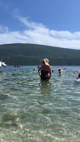 A woman in a wide-brimmed hat and swimsuit stands in the clear, shallow water of a lake or sea. The water is bright and reflects sunlight, with the seabed visible beneath the surface. In the background, other people are in the water. One person is on an inflatable unicorn. Mountains covered in green trees rise from the shoreline in the distance under a blue sky with white clouds.