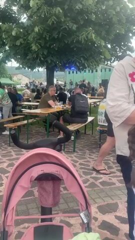 The video shows an outdoor gathering with a stage in the background where a band is performing. Several people are seated at picnic tables, some eating and drinking. In the foreground, a pink baby stroller is visible, and a young girl with braided hair tied with pink ribbons is looking towards the right side of the frame. A woman with short brown hair is walking past the stroller towards the left, looking back over her shoulder. The ground is paved with cobblestones, and a large tree with dense green leaves provides shade. The overall atmosphere appears to be a relaxed outdoor event.