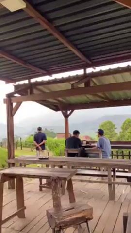 The video shows an open-air wooden structure with a corrugated metal roof, containing several wooden tables and benches. People are sitting and standing around the tables, some interacting with their phones. Through the open side of the structure, a green landscape with trees and distant mountains is visible under a cloudy sky. Some of the vegetation includes banana trees. The camera slowly pans to the right, focusing on the landscape.