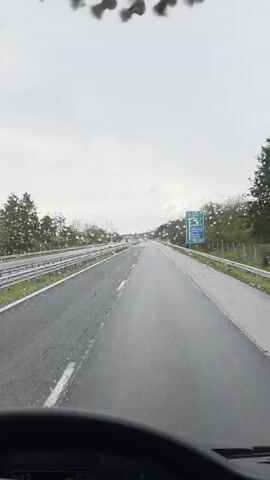 The camera is positioned inside a vehicle driving on a highway under a cloudy sky. The road ahead is straight and mostly empty, with a few other vehicles visible in the distance. The road is wet, with raindrops visible on the windshield. Guardrails run along both sides of the road, with trees lining the background. A road sign displays directions to Varna, Ruse, Tervel, and Tetovo, along with distances. The steering wheel is visible in the foreground, suggesting the viewpoint is from the driver's seat. The windshield wipers are briefly activated at the beginning of the video.