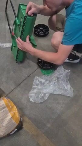 In this media file, a person is assembling a green spreader, likely for lawn care, in what appears to be a garage or workshop. The person is wearing a blue t-shirt and khaki shorts, and is crouched down, holding the spreader with both hands. Two black wheels with green bases are visible on the concrete floor, along with a plastic bag and a bag of what appears to be fertilizer or seed. The lighting suggests it is daytime, and there are no obvious weather cues. The overall mood seems to be one of practical activity, with no strong emotional indicators. The setting is industrial or utilitarian, with a concrete floor and various parts of the spreader scattered around. There is no visible text in the media file.