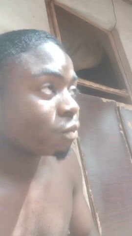 A close-up video shows a shirtless, dark-skinned man with short hair, facial hair, and downturned eyes. He is positioned indoors, against what appears to be a door, possibly made of wood, that has some paint peeling off. A small window or opening is visible at the top of the frame behind the door. The man puckers his lips slightly, as if about to make a kissing sound or whistle. The lighting is somewhat muted.