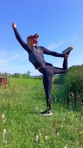The video shows a woman with red hair pulled back in a bun, wearing black leggings and a gray long-sleeved athletic top. She is performing a yoga pose called Natarajasana, or Lord of the Dance pose, in a grassy field under a clear blue sky. Her left leg is extended behind her, bent at the knee, with her left hand holding her left foot. Her right arm is extended upwards. The background features green grass, some flowering weeds, and a bush on the right side. A wooden structure is partially visible on the left. The video pans slightly as she holds the pose.