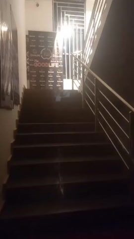 The video shows a dark staircase with a metal railing. There is a poster on the wall that reads "The Goodlife." A bright light shines at the top of the stairs, possibly from a window. The stairs are made of dark material and the walls are painted in light colors. The video is slightly shaky.