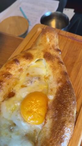 A close-up shot shows a khachapuri, a Georgian cheese-filled bread, served on a wooden board. The bread has a boat-like shape with a golden-brown crust. In the center, a perfectly cooked egg yolk sits atop melted cheese and a white that appears to be slightly undercooked. The lighting is warm and natural, suggesting an indoor setting, possibly a restaurant. The background is blurred but hints at a wooden table and some out-of-focus objects. The overall impression is one of a delicious and freshly prepared dish, ready to be enjoyed.