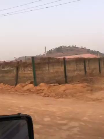 The video shows a vehicle moving along a dirt road past a gas station. The gas station has a white and blue sign displaying the name "NNPC" with the words "RETAIL" underneath. There are tall, thin trees lined up behind the gas station, and a small hill is visible in the background. The sky is overcast. The vehicle appears to be moving at a moderate speed. The view is from inside the vehicle, showing the side mirror on the left.