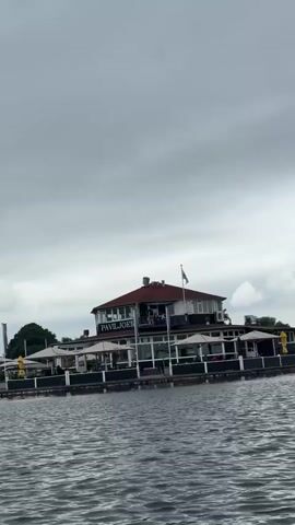 A wide shot captures a building labeled PAVILJOEN on the water's edge. The building has a red roof and a covered outdoor seating area. The sky is cloudy and gray. The water is choppy. Trees and more buildings are visible in the background.