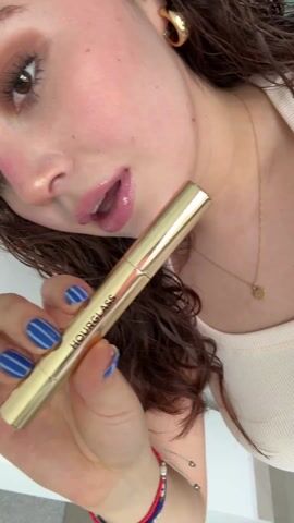 A woman is holding an Hourglass brand lipstick and displays the bottom of the lipstick to the camera. The lipstick is gold in color and says HOURGLASS on the side. The label on the bottom of the lipstick is visible and the lipstick's color, "Crave," is written on it. The woman has brown, wavy hair. She is wearing makeup, including pink lipstick, and has on a gold necklace. She has blue nail polish with white stripes on her nails. A mirror and part of a kitchen can be seen in the background.