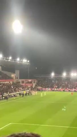 The video shows a soccer game being played at night. A soccer stadium is lit by bright floodlights. The stands are filled with spectators. The players are wearing white and red uniforms. The field is green with white lines. The camera is handheld and slightly shaky.