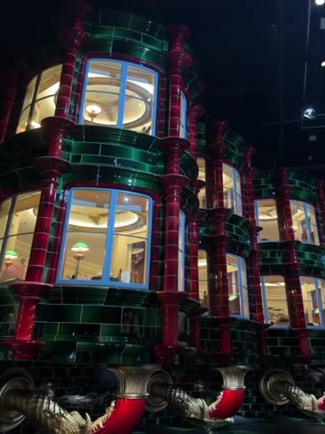The video shows a set built for the Harry Potter movie series, focusing on the exterior of Gringotts Bank. The building has multiple levels of windows and is decorated in green and red tiles with dragon-like accents at the bottom. The interior is lit, indicating activity inside. The camera pans out to reveal a dark, spacious hall with pipes on the ceiling.
