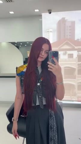 A young woman with long, dark red hair is taking a selfie in what appears to be a salon. She is wearing a grey t-shirt, a dark vest adorned with colorful patterns on the shoulders, and a dark pleated skirt with a patterned sash. She is holding a smartphone in her right hand, angled to capture her reflection. Her left hand is touching her hair. A black salon chair is partially visible to her left. The background shows a large window with a view of buildings, and interior lighting fixtures are visible on the ceiling. The overall scene suggests a moment of self-appreciation or documenting a new hairstyle.