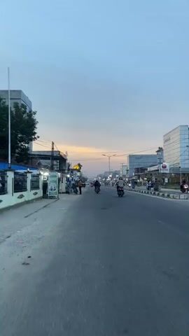 The video shows a street scene at dusk or dawn. The sky is a pale blue with hints of orange and yellow near the horizon, suggesting the sun is either rising or setting. The road is paved and appears to be a multi-lane highway or main road. On the left side of the road, there are several low-rise buildings, some with signs and awnings, and a few trees. A man in a yellow shirt and a man in a blue shirt are standing next to a parked motorcycle in front of one of the buildings. On the right side of the road, there are larger, modern-looking buildings, including what appears to be a supermarket or department store with a large white facade. Several people are visible on the sidewalks and riding motorcycles on the road. The overall impression is of a semi-urban environment transitioning from day to night or vice-versa. There is a sense of activity with vehicles moving and people present.