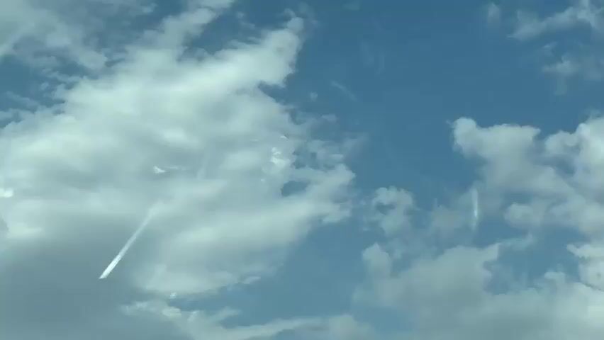 The video shows a clear blue sky with scattered white clouds. There are streaks of light across the sky, possibly caused by reflections or light rays. The clouds are puffy and appear to be moving slowly. The camera appears to be looking upwards, possibly from the ground or from inside a vehicle. The overall impression is of a bright, sunny day.