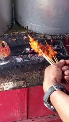 A person wearing a black watch places five skewers of orange-glazed meat over hot coals in a metal grill. The grill is on top of a red wooden stand. A brown, irregularly shaped object sits to the left of the skewers. Two large, round metal containers are visible in the background.