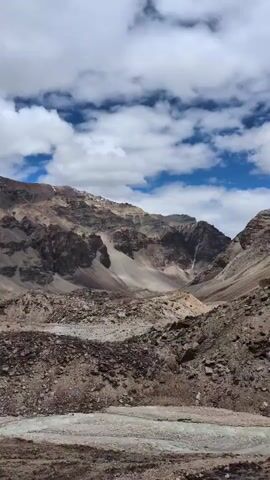 A video pans across a mountainous landscape with a river in the foreground. The mountains are rocky and arid, with patches of snow visible on the higher peaks. The sky is blue with scattered white clouds. The river appears to be shallow and rocky, with a moderate flow. The overall impression is of a remote and rugged environment.
