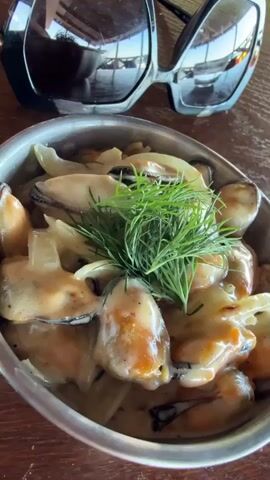 A close-up shot shows a dish of mussels in a creamy sauce, garnished with dill, placed on a wooden table. Next to the dish are a pair of large sunglasses. The camera pans to the right, revealing a view of lounge chairs on a tiled floor, with a railing and the ocean in the background.