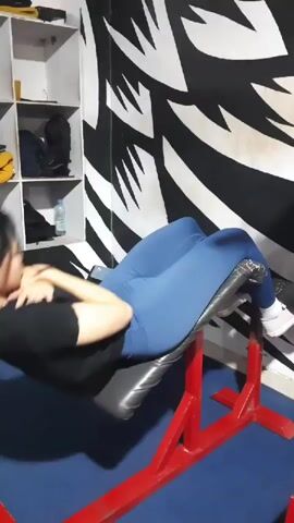 The video shows a woman in athletic wear performing crunches on an angled bench in a gym. The woman is wearing a black t-shirt and blue leggings. Her feet are secured at the top of the bench, and her body is angled downwards. She is seen lifting her upper body towards her knees and then lowering it back down. The gym environment includes shelves with equipment in the background and a black and white graphic design on the wall. The lighting is adequate, allowing for clear visibility of the action. The video is a short loop of the exercise.