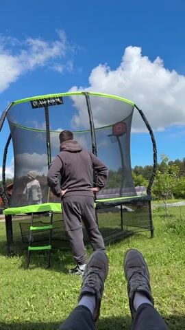 From a low-angle perspective, looking up, a man is seen standing in front of a trampoline on a sunny day. The trampoline is black and green with a safety net and a small basketball hoop inside. A small child is jumping on the trampoline. The man is wearing a gray hoodie and sweatpants with white sneakers. He has his hands on his hips and appears to be watching the child. The person recording the video is lying on the grass, their legs visible in the foreground. The sky is blue with scattered white clouds. In the background, there is a house, trees, and a green lawn. The trampoline has a logo that reads "JUMP POWER".