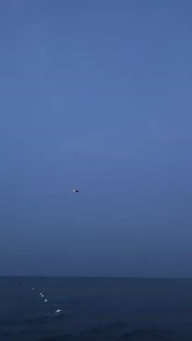 A helicopter flies over a calm ocean during dusk. The sky is a deep blue, transitioning to a lighter shade near the horizon. The water is dark and reflects the dim light. In the distance, along the coastline, several lights are visible, suggesting a populated area. The helicopter is small in the frame, its presence indicated by a faint light.