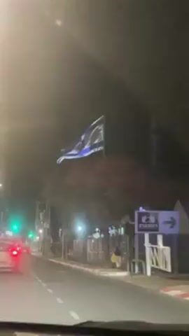 The video shows a car driving at night through a city street, approaching an Israeli flag blowing in the wind. The flag is illuminated by streetlights and has the classic blue and white design with the Star of David in the center.  There are cars on the road, streetlights illuminating the scene, trees lining the road, and a bus stop with a person standing near it. The sky is dark. The perspective is from inside the car, looking out the windshield.  A green traffic light is visible in the distance.