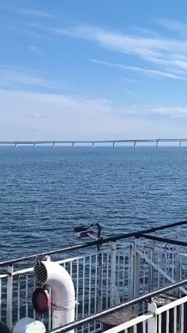 The video shows a wide shot of a calm blue sea under a clear blue sky with scattered white clouds. In the distance, a long, multi-span bridge stretches across the water. In the foreground, the railing and part of a white pipe structure, likely from a ship or ferry, are visible. The water appears slightly choppy with small waves. The overall scene is serene and vast, dominated by the expanse of the sea and sky, with the bridge serving as a significant man-made element in the landscape.