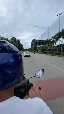 A first-person perspective video shows a person riding a motorcycle on a paved road. The rider, wearing a blue helmet and a white t-shirt, is holding the handlebars. In front of the motorcycle, a white car is driving down the road. To the right of the road, there is a large building with a golf net structure behind it. Palm trees and other greenery are visible along the roadside. The sky is overcast, suggesting cloudy weather. The video captures the motion of the motorcycle as it travels along the road, with the scenery passing by. Another motorcycle is visible further ahead on the right side of the road.