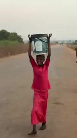 The video shows two young African girls walking on a road, each carrying a bucket on their head. The first girl is wearing a red outfit and carrying a white bucket with a green and blue label on it. She smiles at the camera. The second girl is wearing a yellow shirt and carrying a yellow bucket with other containers stacked on top. The road is unpaved and lined with dry vegetation. There are utility poles in the distance. The sky is overcast.