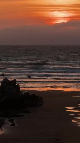The video captures a dramatic sunset over the ocean. The sky is ablaze with vibrant orange and red hues, with clouds partially obscuring the setting sun. The ocean is dark and choppy, with waves rolling towards the shore. A dark, rocky outcrop is visible on the left side of the frame, contrasting with the sandy beach in the foreground. Puddles of water on the beach reflect the colors of the sunset. The overall mood is serene and awe-inspiring.