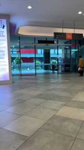 A person in a yellow long-sleeved shirt and black pants walks from right to left across a tiled floor. They are carrying a white bag with a red handle in their right hand. Another person in a red top and blue pants is visible in the background, closer to the glass doors. Further in the background, through the glass, there are buildings with colorful stripes. The scene appears to be an indoor walkway or mall.