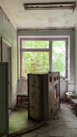 The video shows the interior of a dilapidated room. The walls have peeling paint and visible water damage. There is a large, rusted metal control panel-like object in the center of the room, with a window behind it revealing a view of dense green trees outside. The floor is covered in debris and broken tiles. There is a stool or small table to the left and right of the control panel. In the background, an open doorway reveals another room, similarly damaged, with debris scattered across the floor and another window providing a view of the trees. The lighting is dim due to the state of the room and the light filtered through the trees.