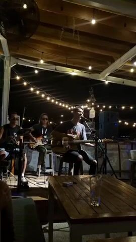 Here's a description of the video:

Three musicians are performing live on a small stage in an outdoor setting at night. They are under a partially covered patio or roof adorned with string lights. The vocalist, seated in the center, is playing an acoustic guitar and singing into a microphone. To his left, a guitarist sits on a stool, playing an electric guitar. On the far left, a drummer is playing a basic drum kit.

A small table with a wooden top is in the foreground, with a half-empty glass of beer or another beverage. The space appears to be a bar or restaurant with other patrons visible in the background. A large fan is visible on the ceiling. The ambiance is relaxed and casual.