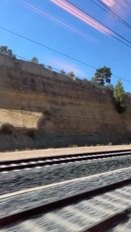 The video shows a view from a moving train, looking out the window. The train tracks are visible in the foreground, with ballast and ties. Beyond the tracks, there is a sloped embankment covered in dry vegetation and a few trees. The embankment is rocky with some sandy areas. A cut in the embankment is visible, suggesting a possible tunnel or construction. The sky is clear and blue, with a few power lines running horizontally across the frame. The reflection of the train's interior lights is visible on the window, creating streaks of light. The overall impression is of a sunny day in a dry, hilly landscape.