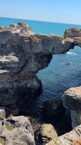 The video displays a rocky coastline with the turquoise sea. Several people are present on the cliff tops, some standing and observing the view, others seated. The cliffs are rugged and appear to be made of sedimentary rock. The water is clear, and the sky is a bright blue with no clouds visible. The video pans across the coastline, showing the formation of natural arches and sea stacks. There are also rocks scattered in the water near the shore. The overall impression is of a scenic coastal environment on a sunny day.