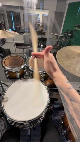 The video presents a drummer's viewpoint from behind a drum set in a studio setting. The drummer's left forearm, tattooed with an image of a man and a car, is visible as the drummer performs stick tricks with a drumstick. A drum set, comprised of several drums of various sizes, with a white-headed snare drum positioned prominently in the foreground, is evident. The drums are predominantly brown with silver hardware. A cymbal is partially visible on the right side of the frame. The recording space is equipped with microphones and soundproofing material visible along the walls. A mirrored surface in the background reflects another person, who appears to be monitoring the recording session.