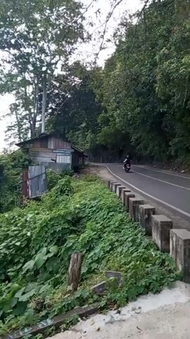 The video begins with a shot of a small, rustic wooden shack nestled on a steep, overgrown hillside beside a paved road. A motorcycle is seen riding away from the camera on the road. The camera then pans to the right, revealing a panoramic view of a lush green coastline meeting a calm turquoise ocean under a cloudy sky. In the distance, several small islands are visible. The scene appears to be a remote, natural landscape.