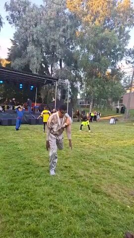 A young Black male wearing a shiny silver jumpsuit and white sneakers is performing a dance routine on a grassy lawn. He is facing away from the camera with his left leg kicked forward and his arms extended outwards. In the background, there is a stage with performers, trees, a building, and an audience.