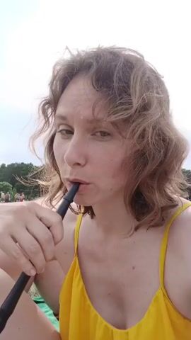 A woman with curly brown hair, wearing a yellow tank top, sits outdoors on a grassy area, holding a black pipe-like object to her mouth and exhaling a cloud of smoke. The background shows a body of water, trees, and a cloudy sky.
