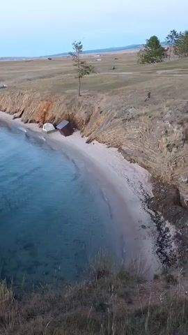A vertical video shot from a high vantage point overlooking a tranquil bay. The water is a clear, deep blue, transitioning to lighter turquoise near the shore. A sandy beach curves along the coastline, meeting a gentle slope of dry grass and rocky outcrops. On the beach, nestled against a cliff face, sits a small, rustic, red-roofed structure, partially obscured by a white tent. Further up the beach, a lone tree stands on the grassy plateau. In the distance, the landscape stretches into rolling hills under a pale, overcast sky. The scene is serene and shows no human activity.