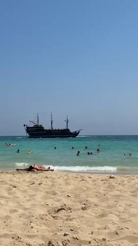 The video shows a beach scene with a pirate ship in the distance. In the foreground, there is a sandy beach. In the middle ground, there is turquoise water with several people swimming and sunbathing. One person is lying on the sand in the foreground. The sky is clear and blue.