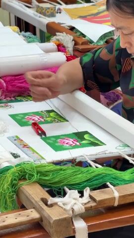 In a close-up shot, a woman, likely an embroiderer, is meticulously working on a piece of hand embroidery. She is seated, with her hands deftly guiding a needle and thread through a white fabric stretched taut on a wooden embroidery frame. The frame rests on a wooden stand, and bundles of vibrant green and pink embroidery threads are visible around it.

On the fabric, there are two square sections, each featuring a pre-printed design of a pink lotus flower against a green background. The woman is actively embroidering the green background of the design on the right. A small red thread cutter and a ruler are placed on the fabric near her working area. Other embroidery supplies and spools of thread are visible in the background, suggesting an indoor workshop or studio setting, consistent with craft activities in Hải Phòng, Vietnam. The woman is focused and engrossed in her detailed work.